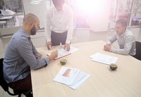 Drei Männer im Besprechungsraum. Zwei Männer sitzend und ein Mann stehend während einer Unterhaltung an einem Holzschreibtisch mit Unterlagen und zwei schwarzen Tassen an Kaffee auf dem Tisch.